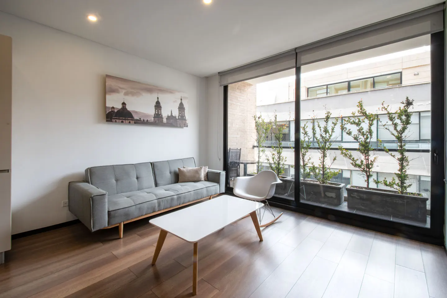 Bright living room with gray sofa, white table, and rocking chair. Balcony with plants visible through large windows. Cityscape art on the wall.