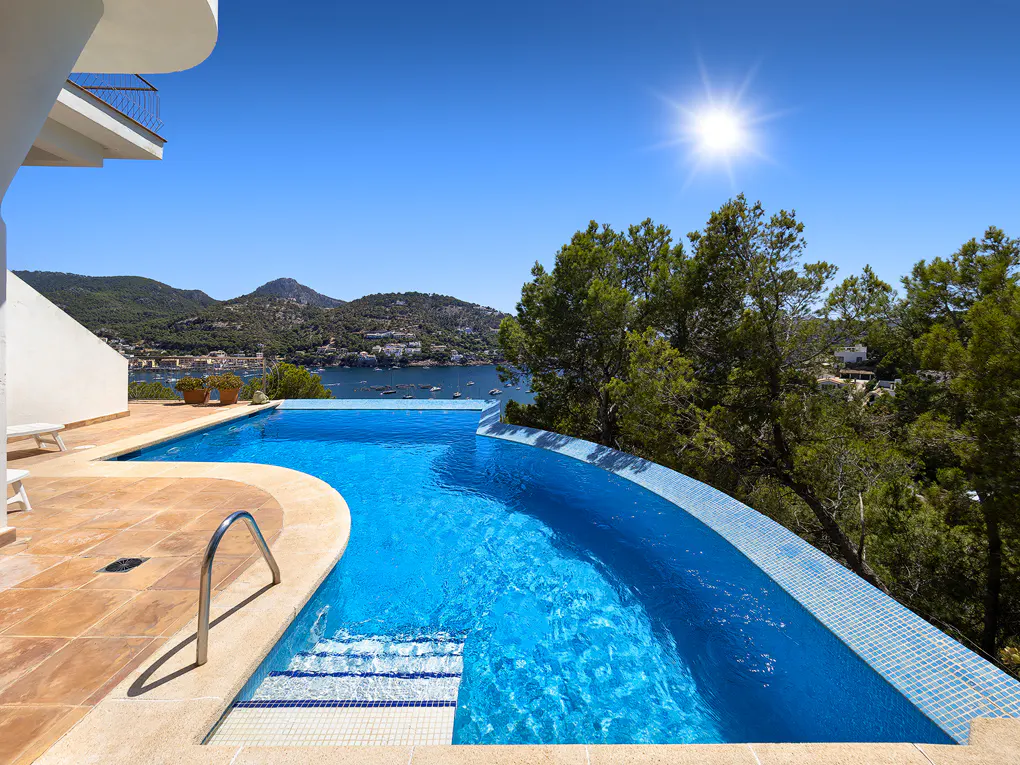 Infinity pool with blue water overlooking a bay with mountains and trees under a bright blue sky.