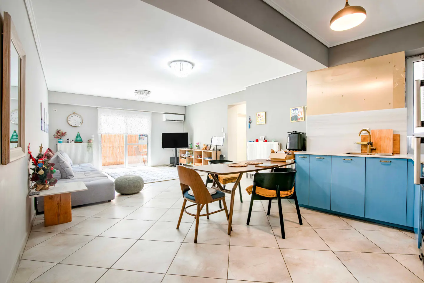 Open-plan living area with blue kitchen cabinets, a dining table, and a gray sofa in a bright, tiled room.