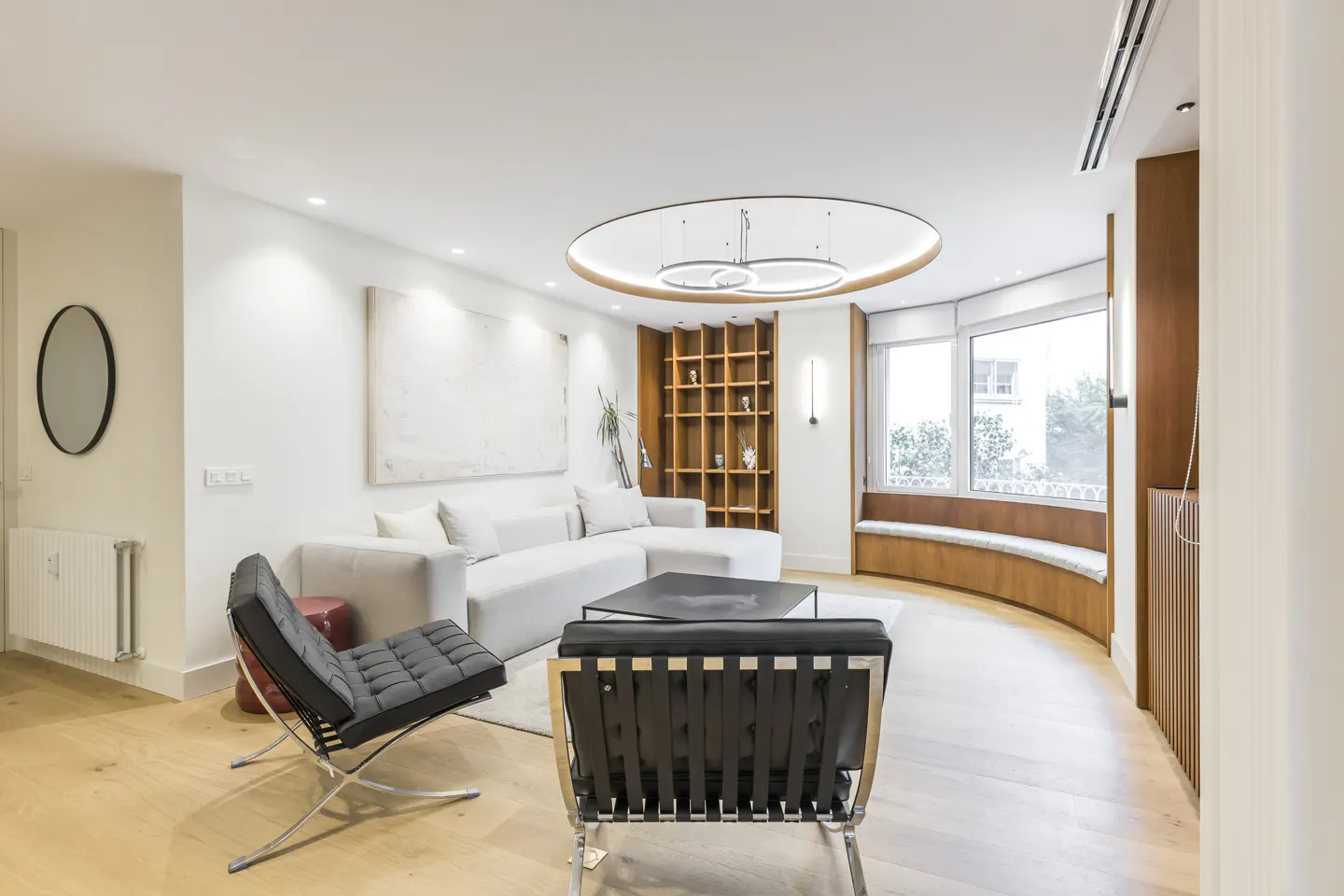 Modern living room with white sectional sofa, two black leather chairs, wooden shelving, and a curved window seat.