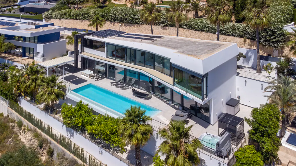 Aerial view of a modern white house with a pool, palm trees, and solar panels on the roof.
