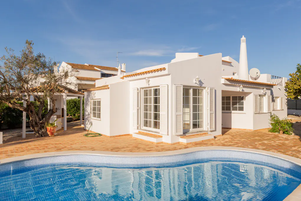 Exterior view of a white villa with a blue tiled pool in the foreground on a sunny day.