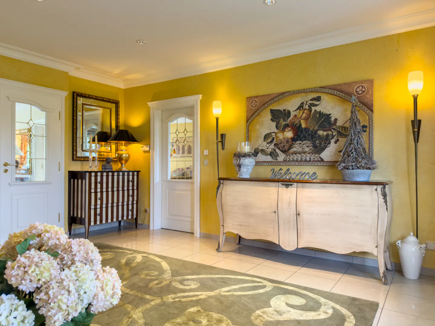 Foyer with yellow walls, tile floor, and a green rug. A white cabinet with a painting above it is on the right. A striped dresser is on the left.