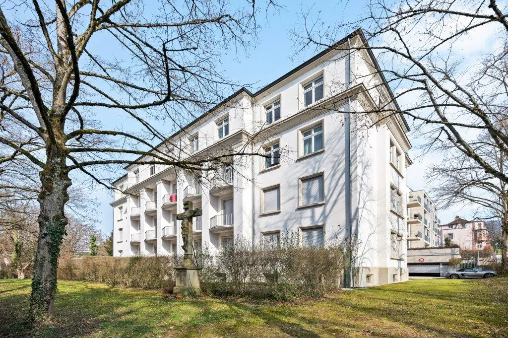 Exterior view of a white apartment building with balconies, a stone cross in the yard, and bare trees against a blue sky.