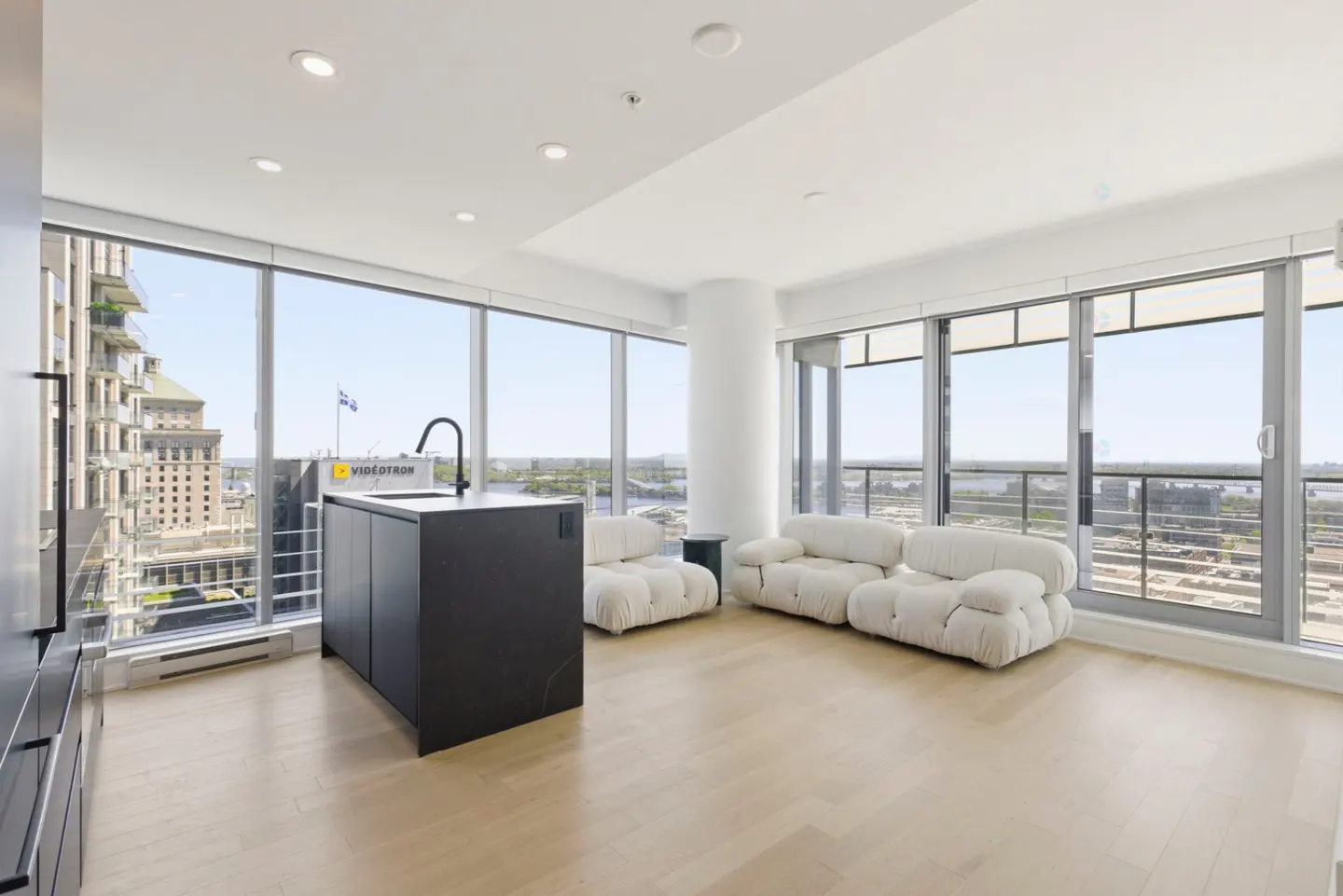 Bright, modern condo with floor-to-ceiling windows, light wood floors, and white sofas. A black kitchen island stands near the window with city views.