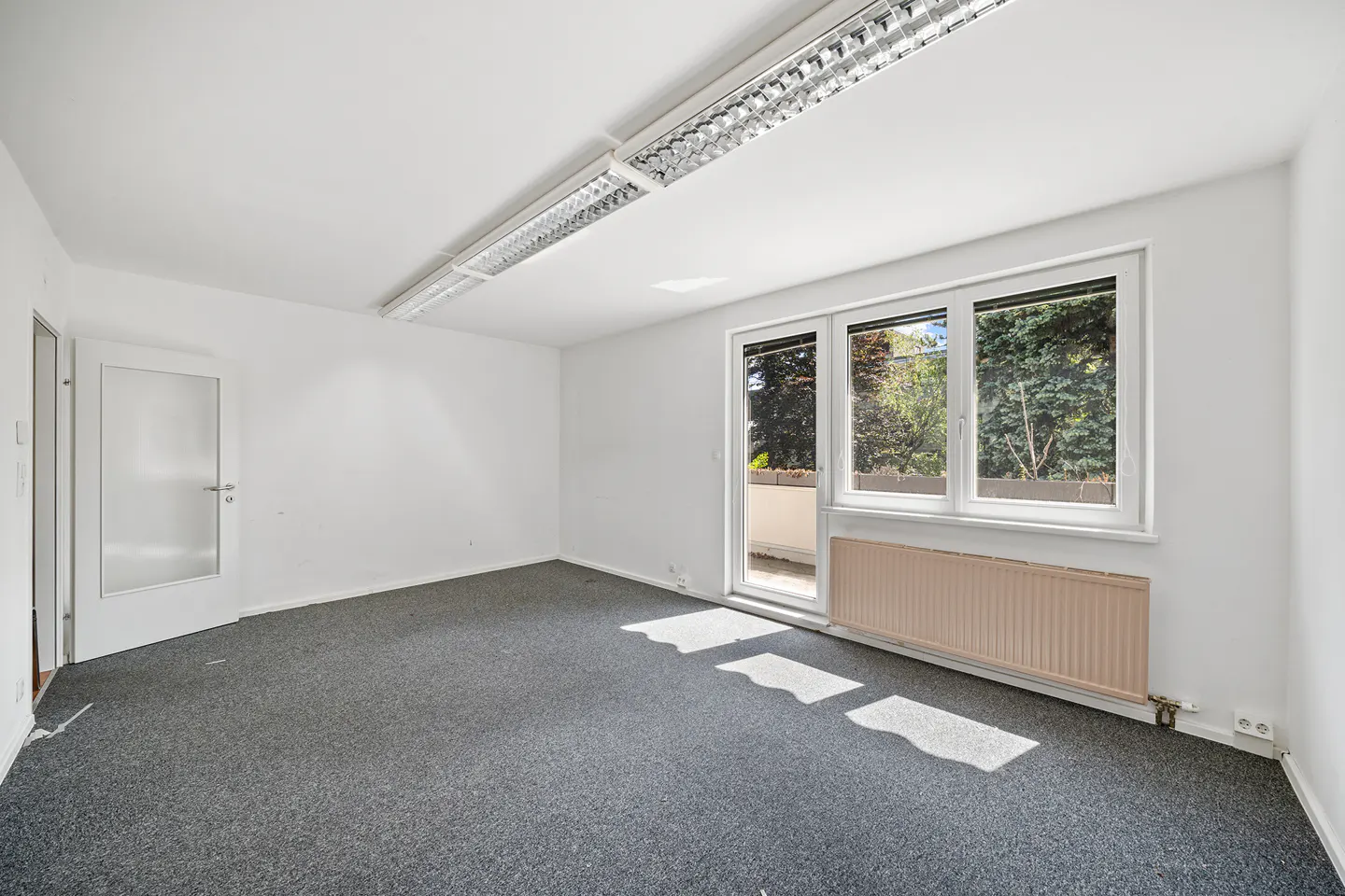 Empty room with white walls, grey carpet, a glass door to a balcony, a window, and a beige radiator under the window.