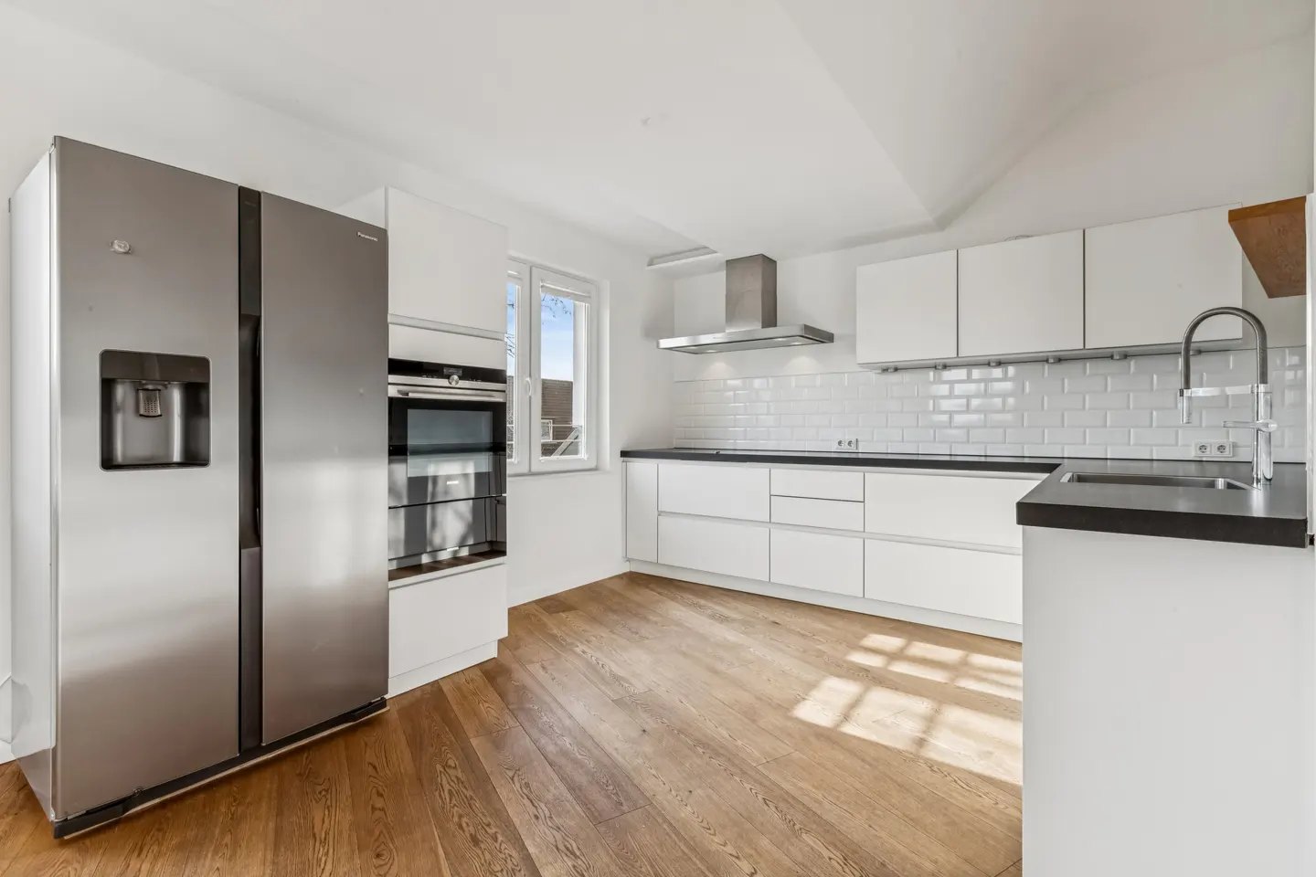 Bright kitchen with wood floors, white cabinets, and black countertops. A stainless steel refrigerator stands to the left.