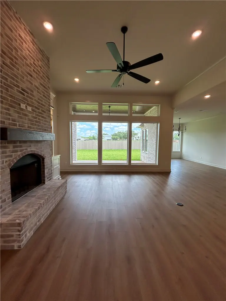 A spacious living room with wood floors, a brick fireplace, and a large window overlooking a green lawn. A ceiling fan hangs above.