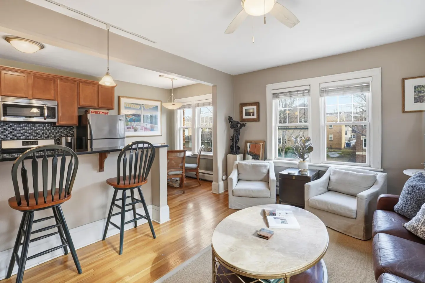 A well-lit living room and kitchen area with hardwood floors, two white armchairs, and a brown leather sofa.