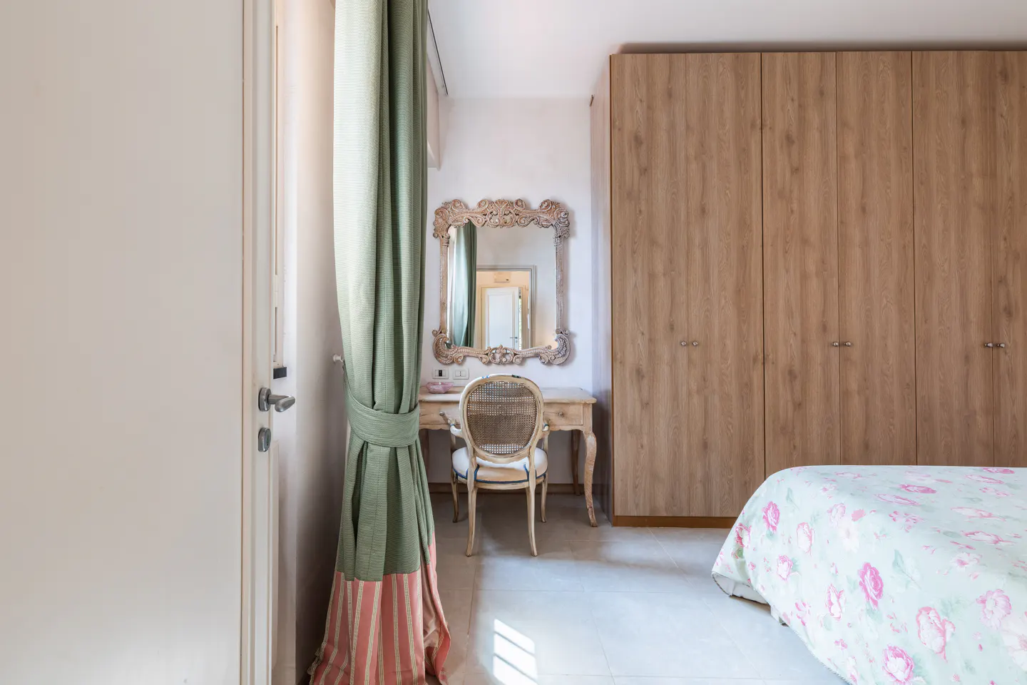 Bedroom with a vanity, mirror, and chair. A green curtain is tied back. A bed with a floral comforter is visible. A wooden wardrobe stands against the wall.