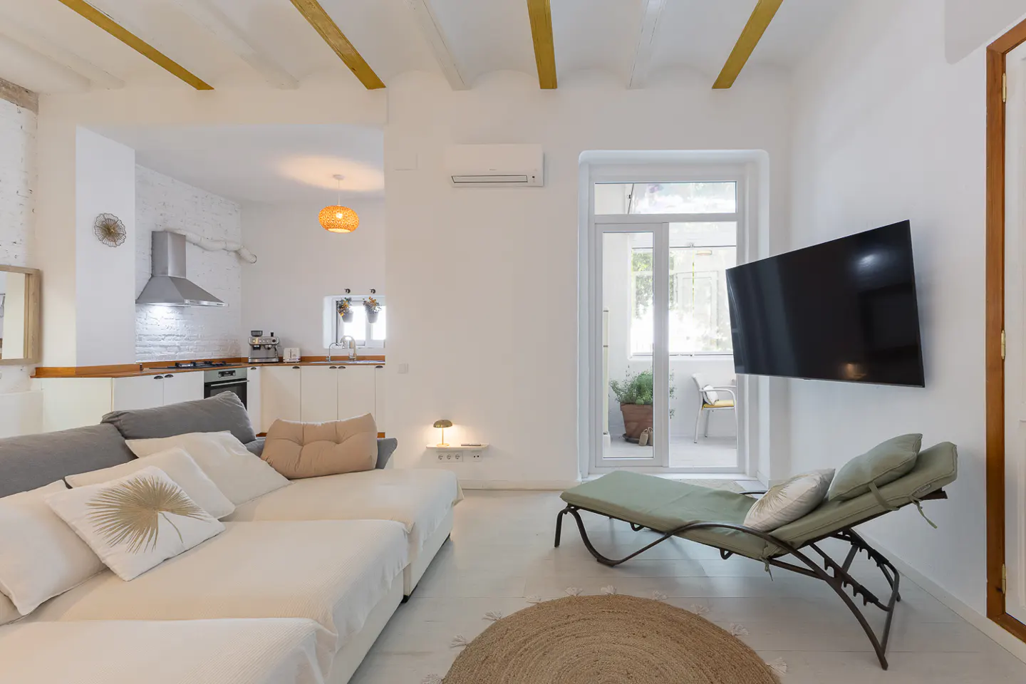 Bright, open-concept living space with white walls, wood beams, and a jute rug. A large sectional sofa faces a TV and a chaise lounge. Kitchen visible in the background.