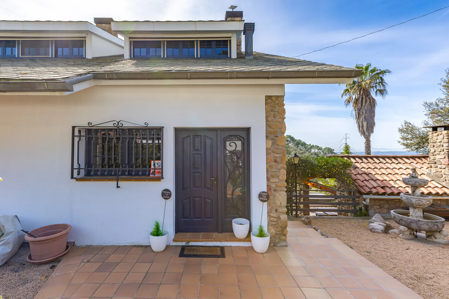 Exterior of a white house with a gray door, stone accents, and a tiled patio. A palm tree is visible in the background.