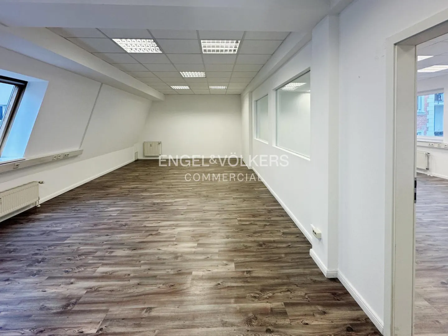 Bright, empty commercial space with wood-look floors, white walls, and recessed lighting. Engel & Völkers Commercial logo visible.