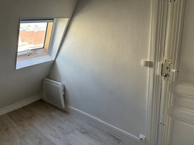 Attic room with white walls, light wood flooring, a slanted ceiling, a small window, and a wall-mounted heater.