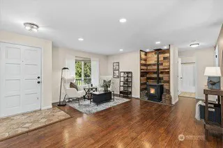Bright living room with hardwood floors, white walls, and a white front door. A wood-burning stove sits in a corner with a wood accent wall. A white chair and black table sit on a gray rug.