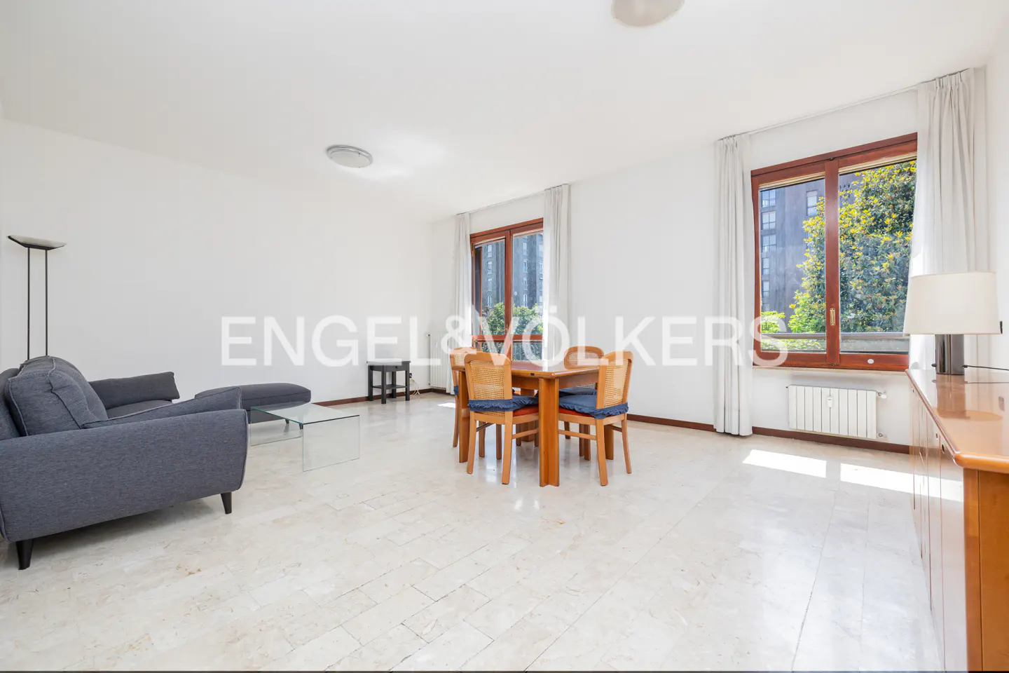 Bright living room with a gray sofa, glass table, wood dining set, and large windows with white curtains.