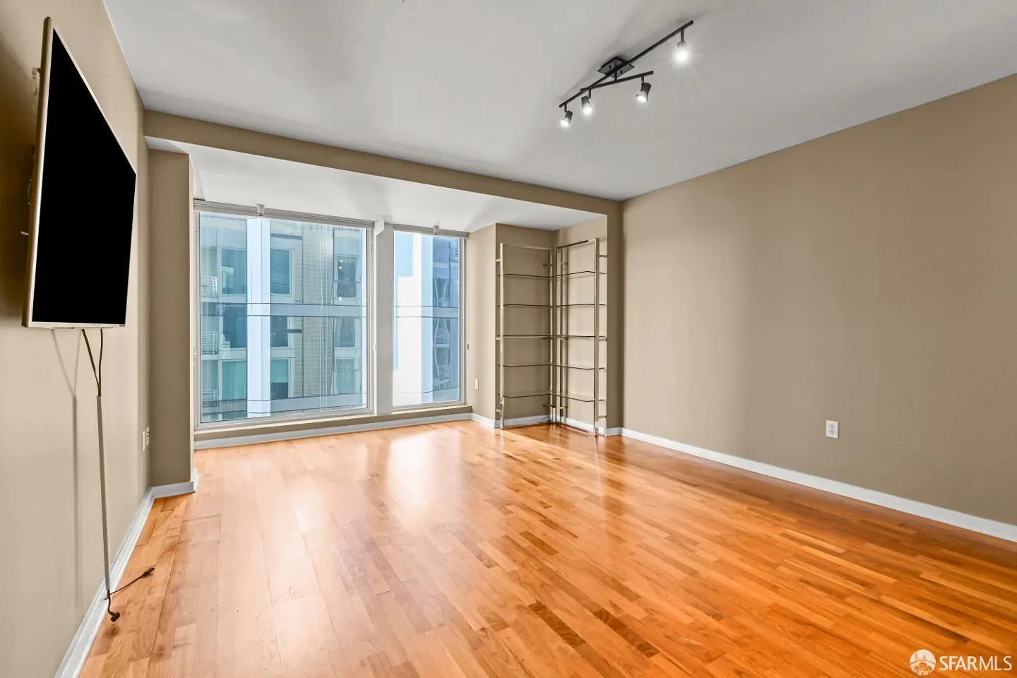 A bright, empty room with hardwood floors, beige walls, and large windows with a city view. A TV is mounted on the wall, and a metal shelving unit stands in the corner.