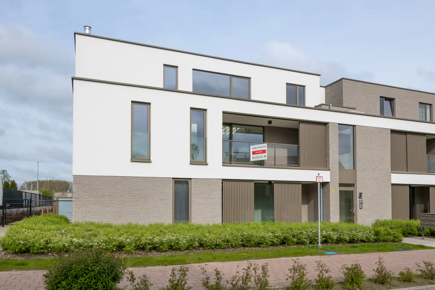 Modern apartment building with white and gray brick facade, balcony with "For Sale" sign, and green landscaping.