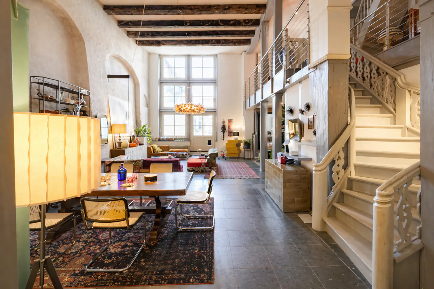 Bright, open living space with high ceilings, exposed beams, and large windows. A wooden table and chairs sit on a dark rug. White staircase on the right.