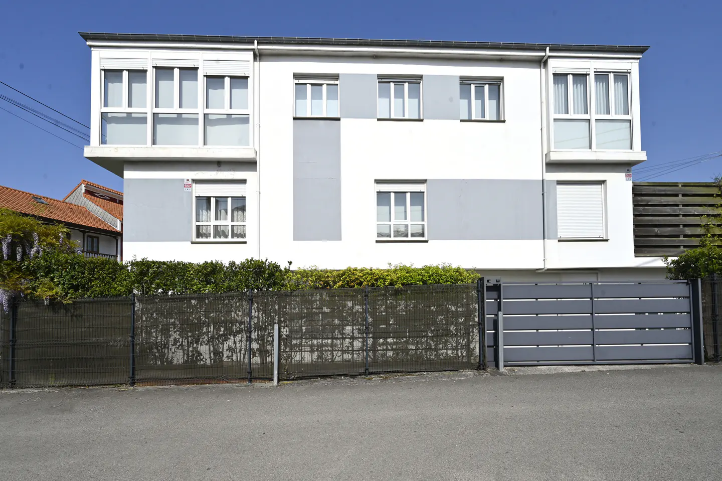 Two-story white house with gray accents, multiple windows, and a gray horizontal slat gate. Green bushes line the fence. Blue sky.