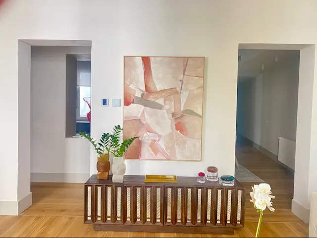 Living room with wood floors, white walls, and a brown cabinet. An abstract painting hangs above the cabinet, with plants and decorative jars on top.