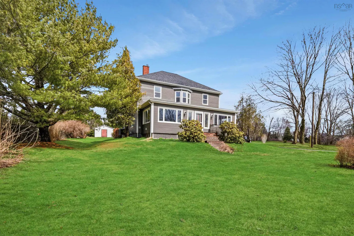 Two-story gray house with a red brick chimney, surrounded by a green lawn and trees under a blue sky.