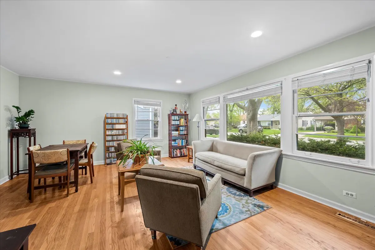 Bright living room with hardwood floors, a beige sofa, an armchair, and a dining table. Large windows offer a view of a green neighborhood.