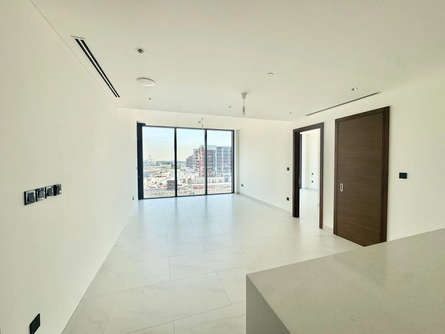 Bright, empty apartment with white walls and tile floors. Large window shows a city view. Brown doors and black switches add contrast.