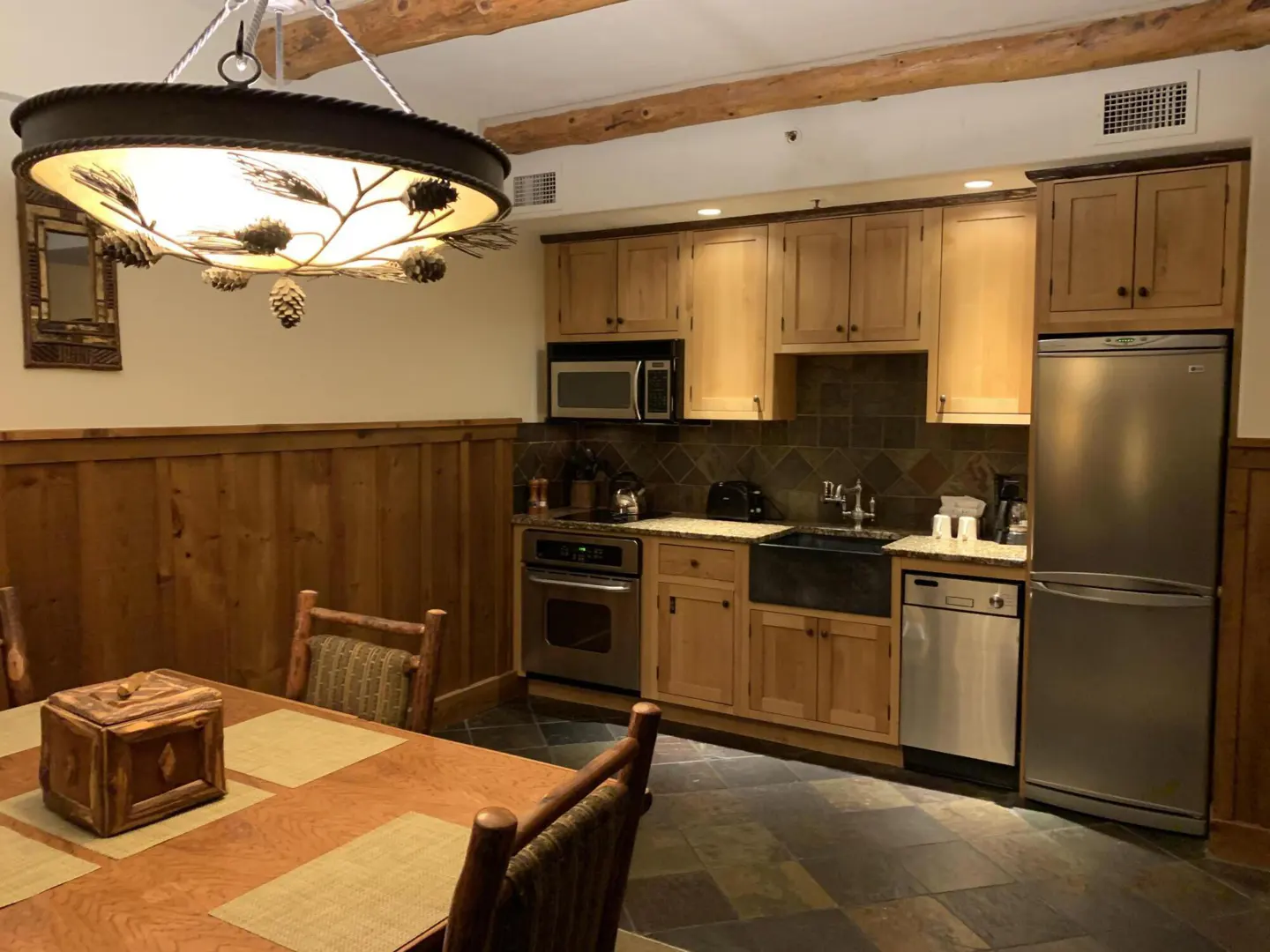 A kitchen and dining area with wood paneling, light wood cabinets, and a pinecone chandelier.