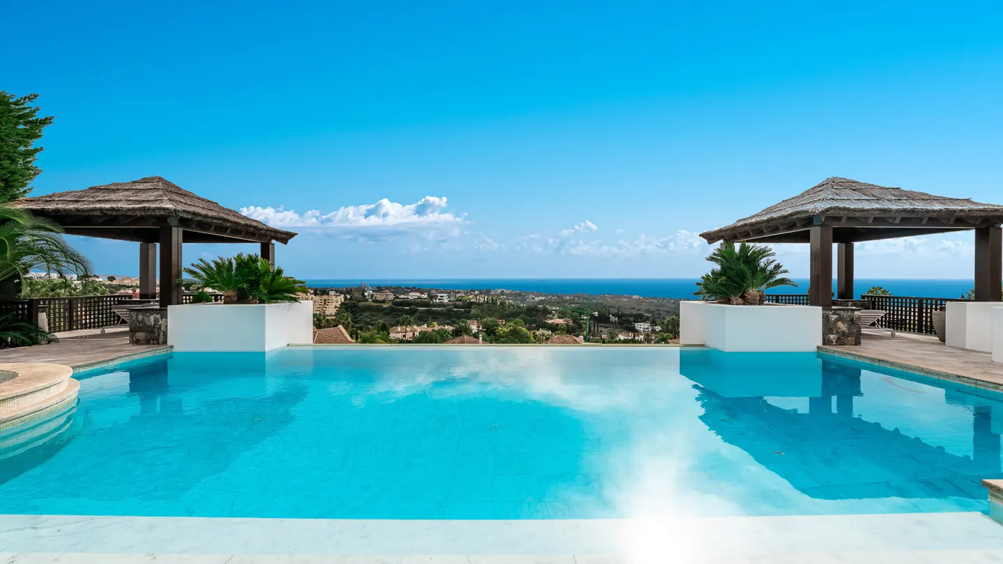 Infinity pool with two thatched-roof gazebos overlooking a coastal landscape under a clear blue sky.