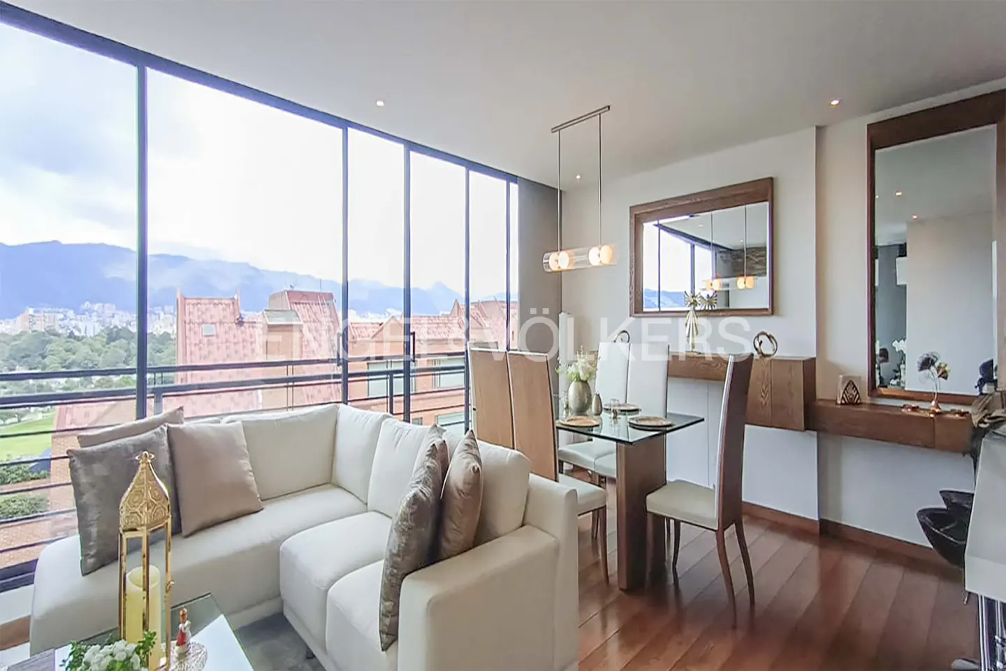 Bright living room with a white sofa, glass dining table, and floor-to-ceiling windows overlooking a city and mountains.