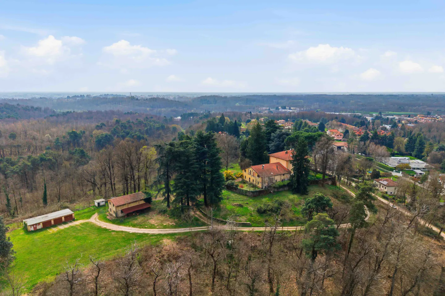 Aerial view of a yellow house with a red tile roof, surrounded by trees and greenery. Blue sky with white clouds above.
