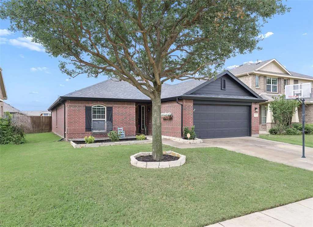 A single-story red brick house with a gray roof and a dark gray garage door. A large tree stands in the front yard.