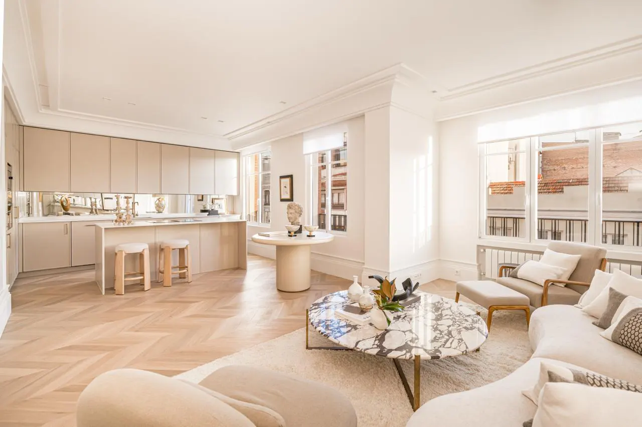 Bright, modern apartment interior with white walls, herringbone wood floors, and beige kitchen cabinets. A marble coffee table sits in front of a curved sofa.