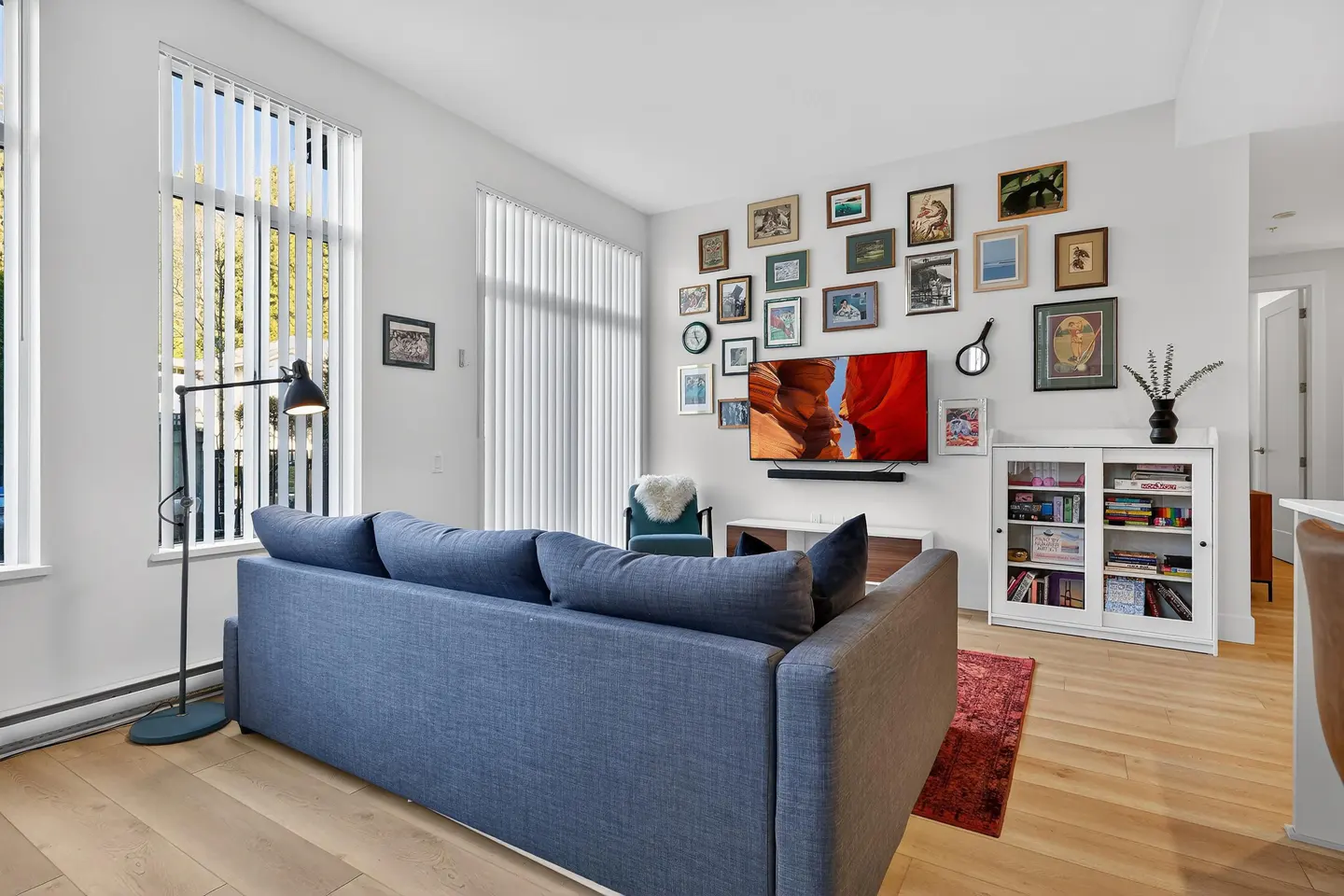 Bright living room with a blue sofa, light wood floors, and a gallery wall with a TV. Tall windows with white blinds let in natural light.