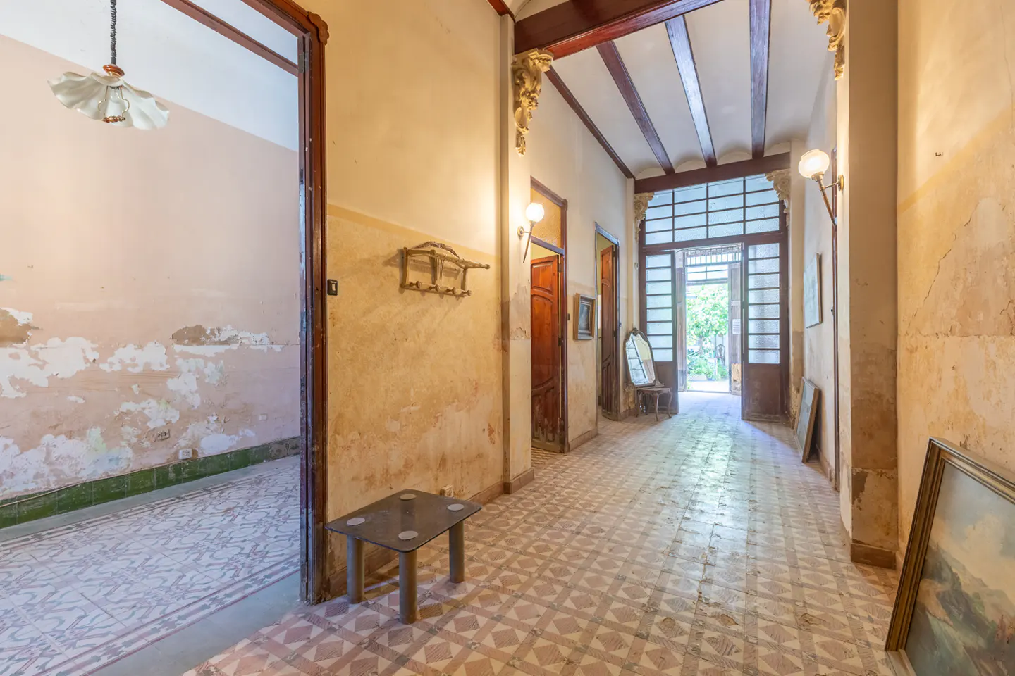 Hallway with patterned tile floor, aged walls, and wooden beams. A table and coat rack are on the left, with an open doorway to a room.
