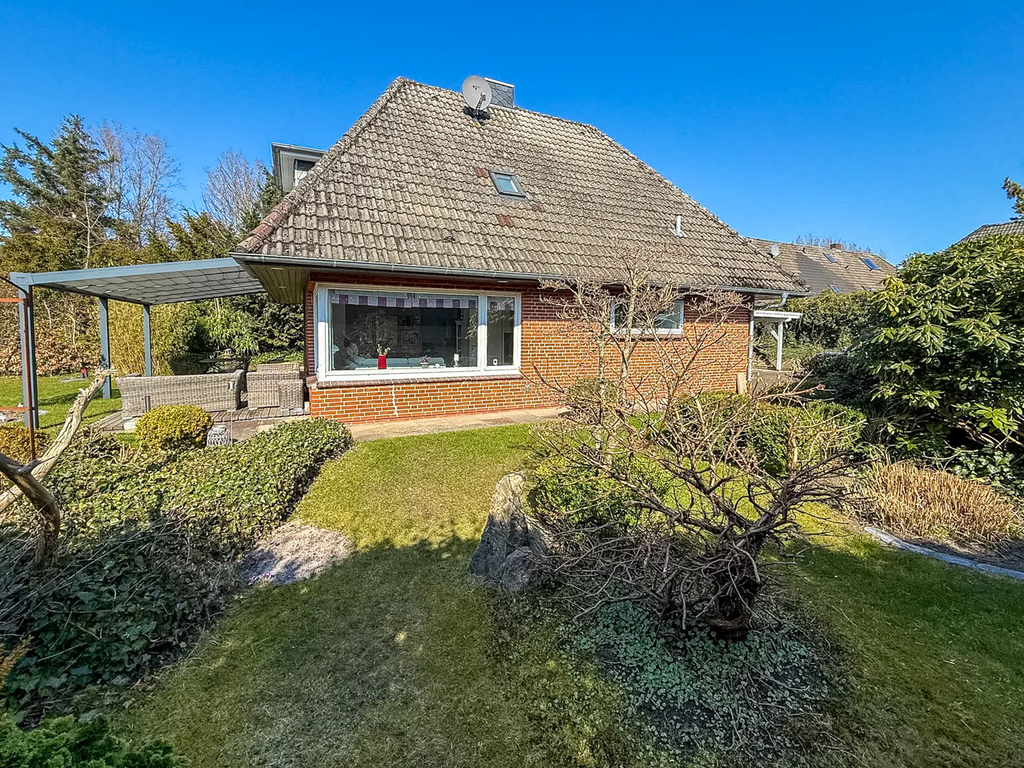 A single-story brick house with a gray tiled roof and a covered patio with wicker furniture on a sunny day.