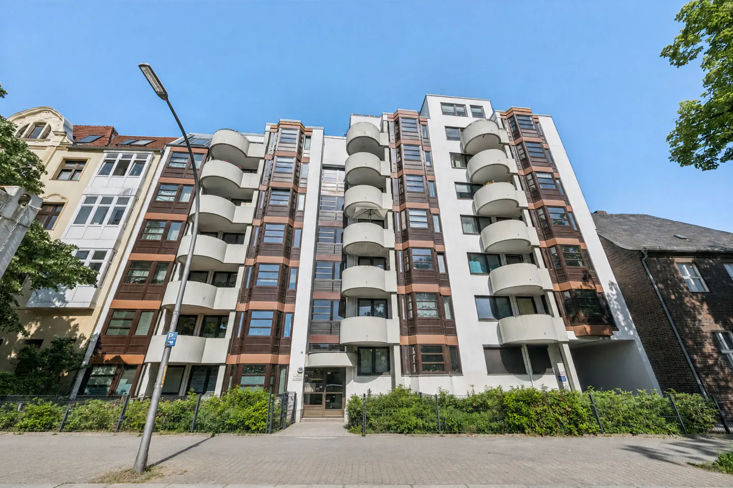 Low-angle view of a modern white apartment building with rounded balconies, brown accents, and a sidewalk in the foreground.