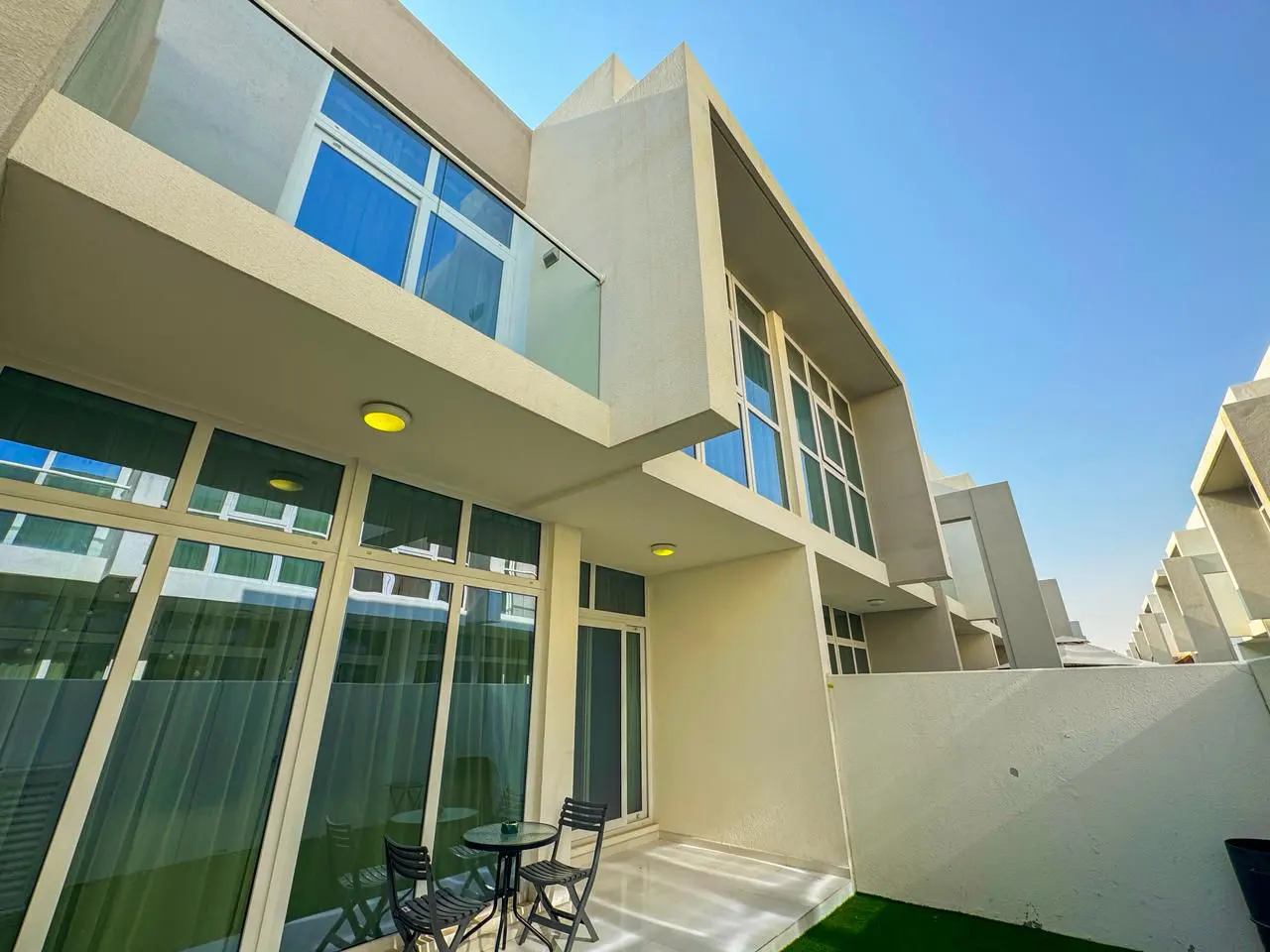Exterior view of a modern, light-beige townhouse with large windows, a small patio with a table and chairs, and a clear blue sky.