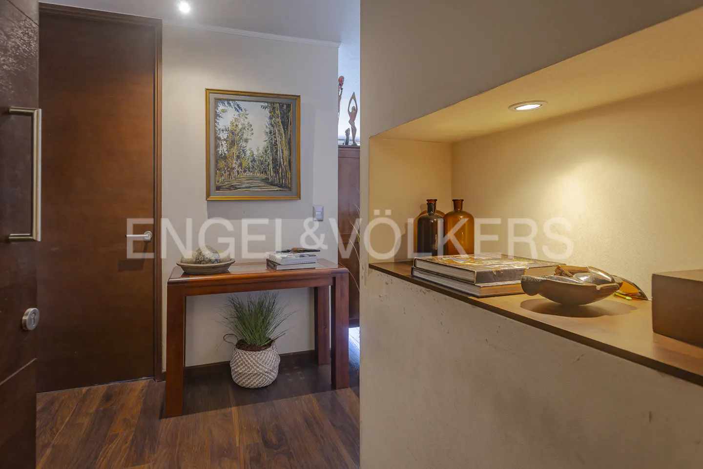 Entryway with a wood door, a framed landscape painting, a table with a plant, and a shelf with books and amber bottles.