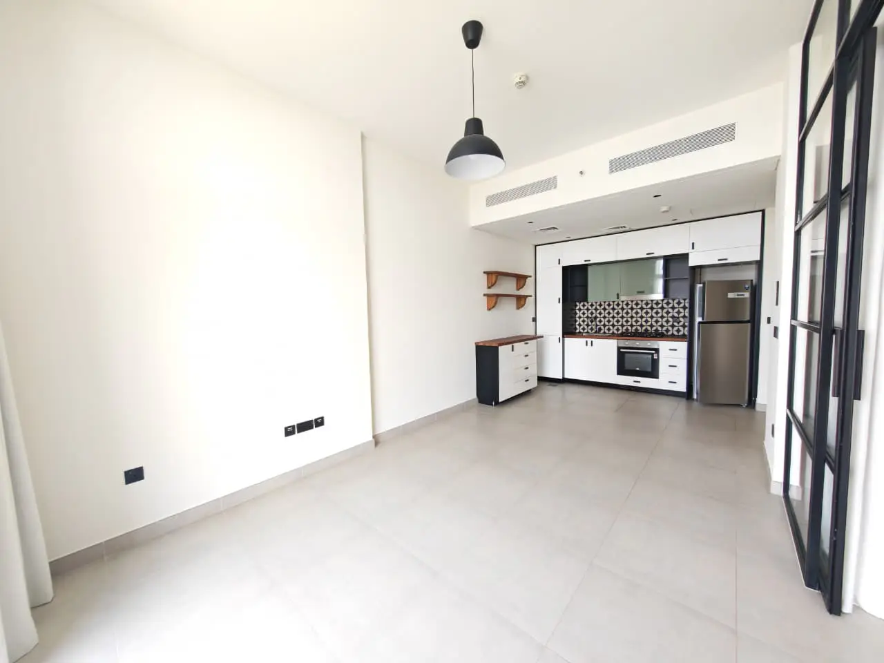 Bright, modern apartment interior with white walls, tile floors, and a black and white kitchen. A black pendant light hangs from the ceiling.