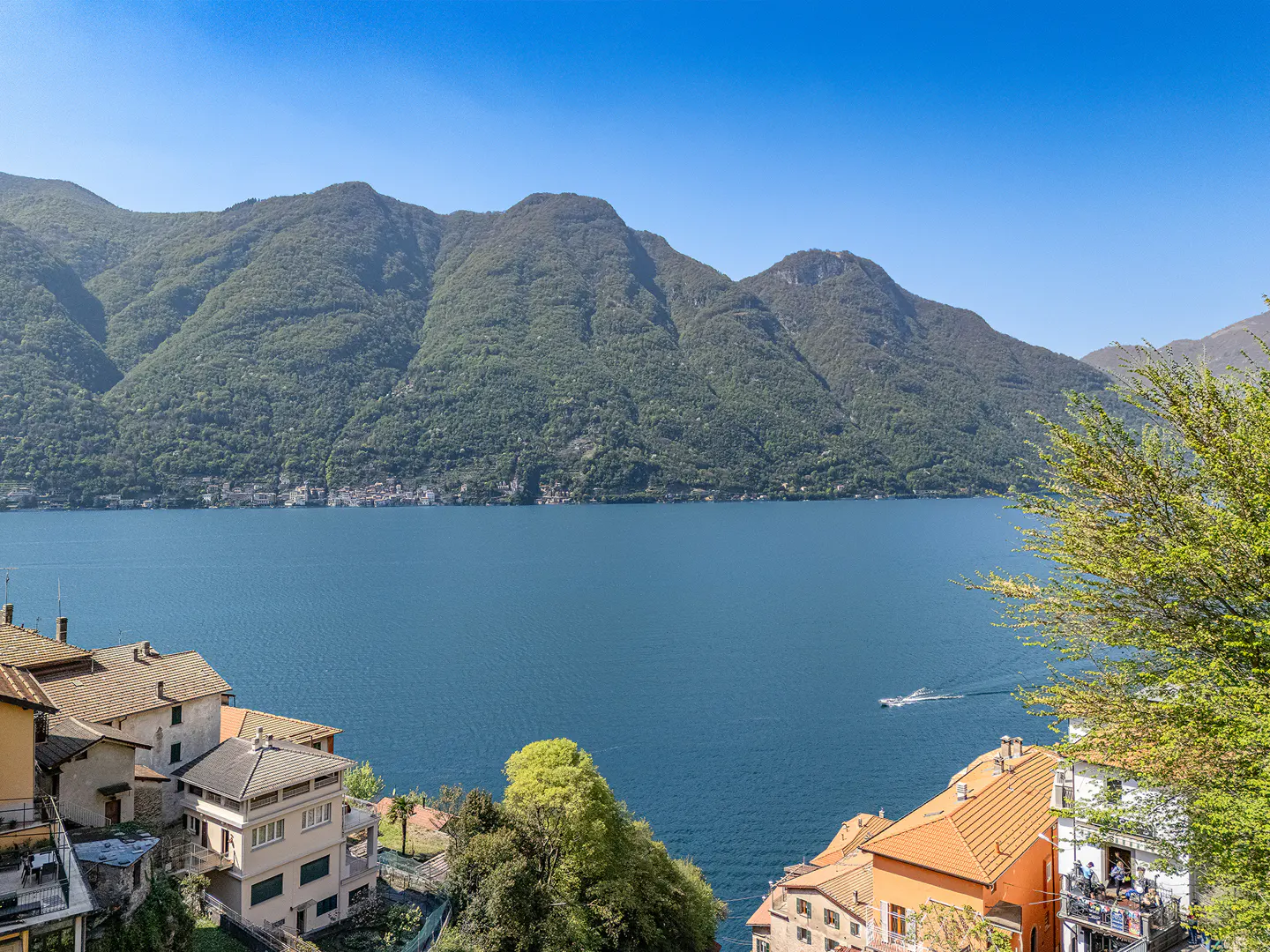 Scenic view of Lake Como, Italy, with mountains, blue water, and colorful houses with orange roofs. A boat is seen on the lake.