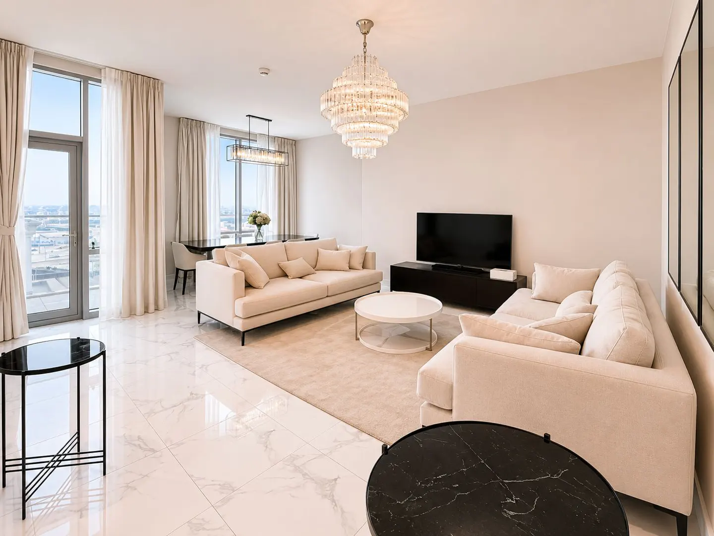 Bright living room with beige sofas, a white rug, and marble floors. A crystal chandelier hangs above, and large windows offer a city view.