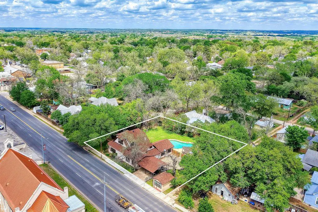 Aerial view of a property with a pool, surrounded by trees and a white border, next to a road and other buildings.