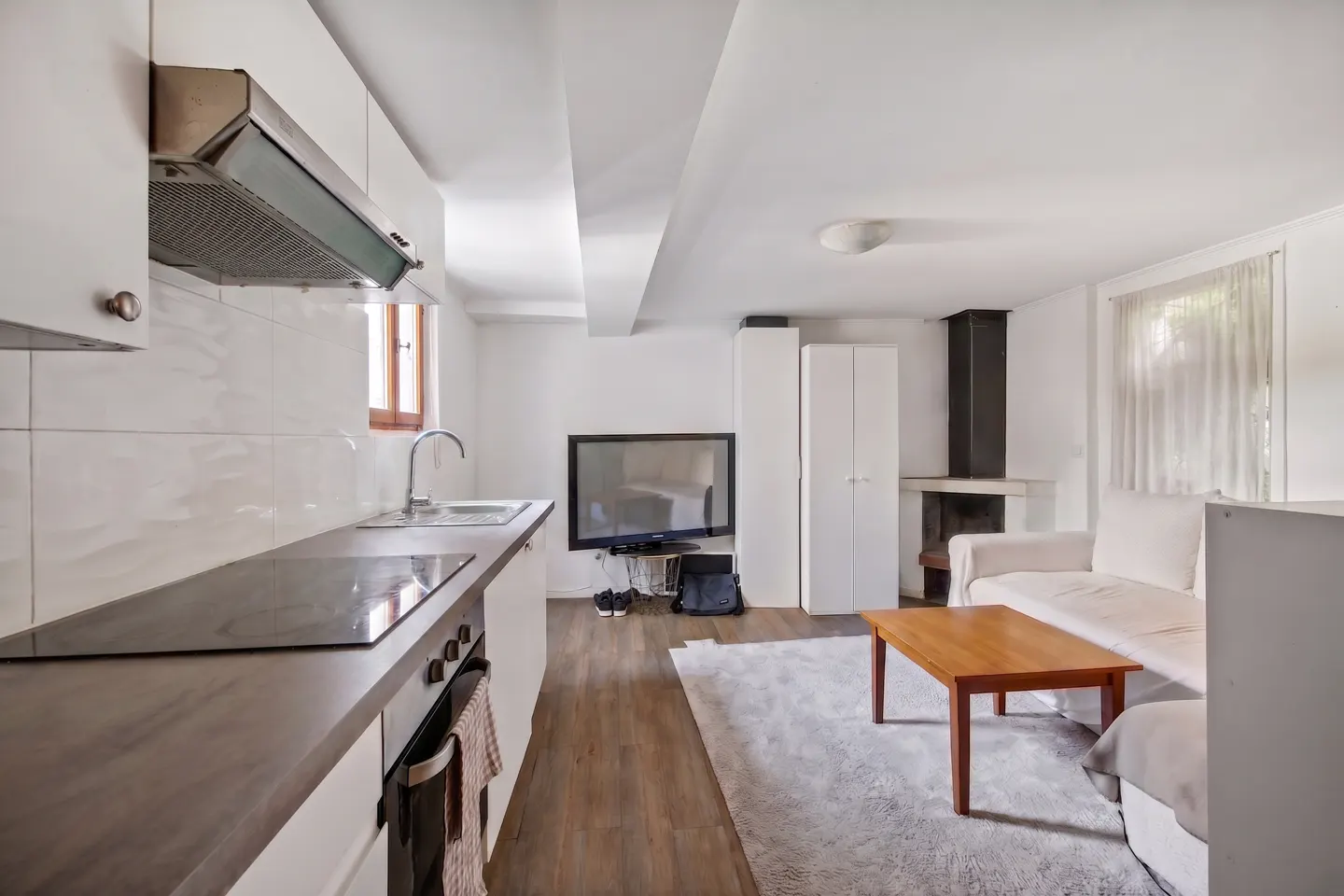 Open-concept apartment with white walls, wood floors, and a kitchen area with gray countertops. A living area has a white sofa, wood table, and fireplace.