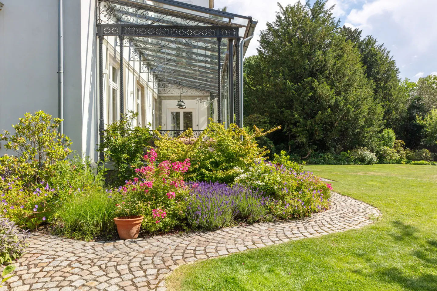 A garden path curves past a flowerbed with pink and purple flowers, leading to a glass-roofed veranda attached to a white house.