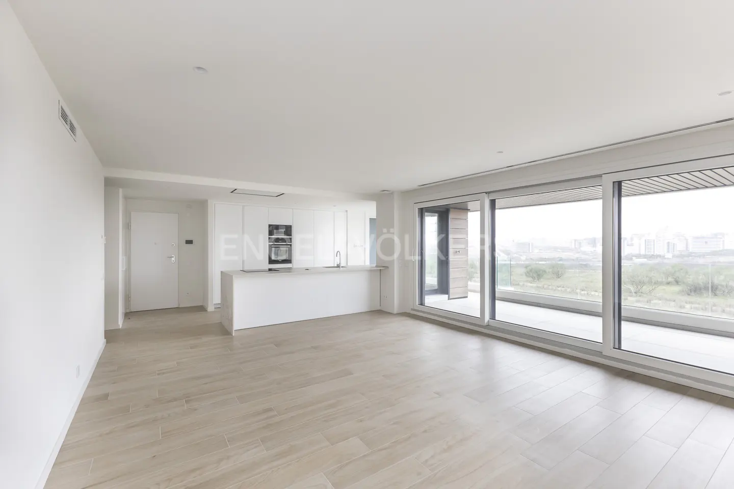 Bright, modern apartment with light wood floors, white walls, and a kitchen island. Large sliding glass doors open to a balcony with a city view.