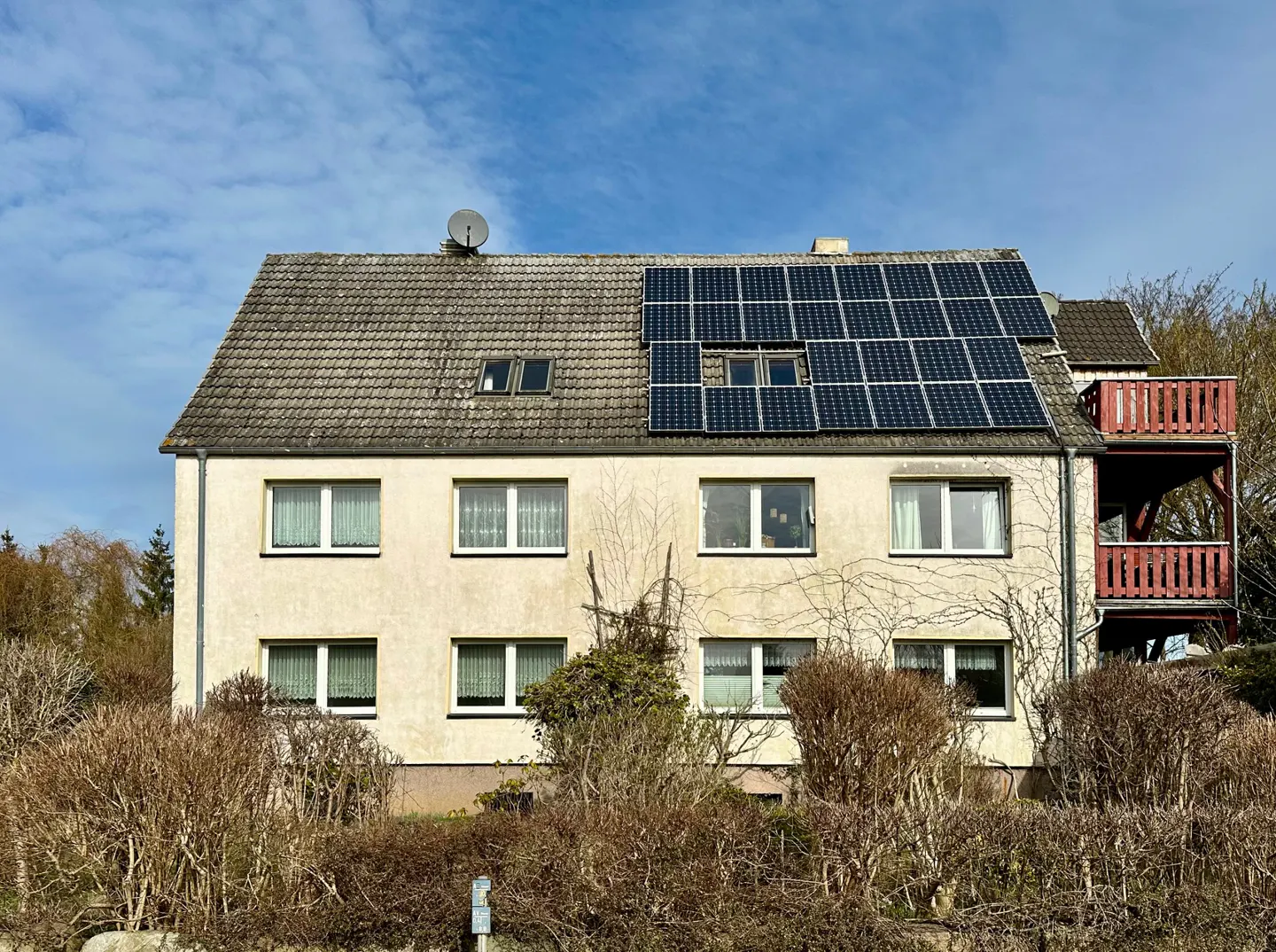 Two-story beige house with solar panels on the roof, a red balcony, and a satellite dish against a blue sky.