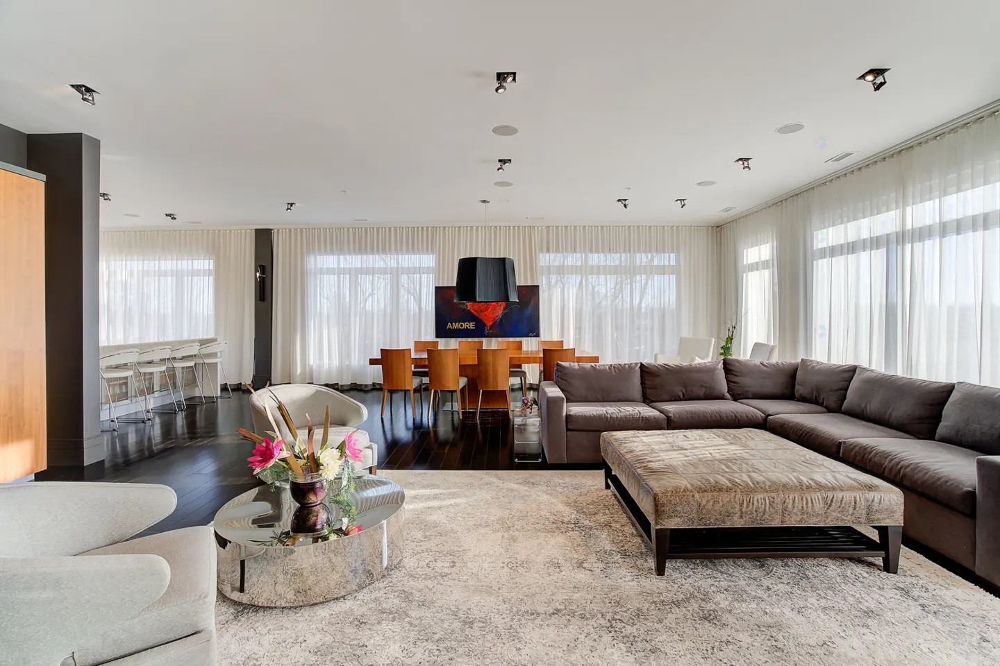 Bright, open-concept living room with dark wood floors, a large gray sectional, and a dining table with "AMORE" art. White sheer curtains.