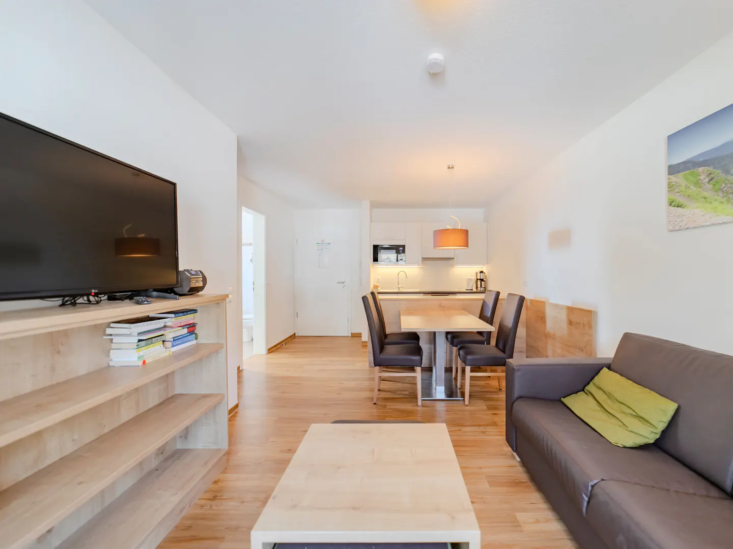 Bright apartment interior with wood floors, a gray sofa with a green pillow, a dining table, and a kitchen in the background.